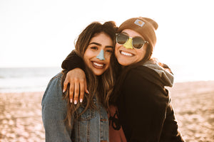 Two smiling enthusiastic young women. Hugging on the beach wearing colorful and vibrant nose sunscreen